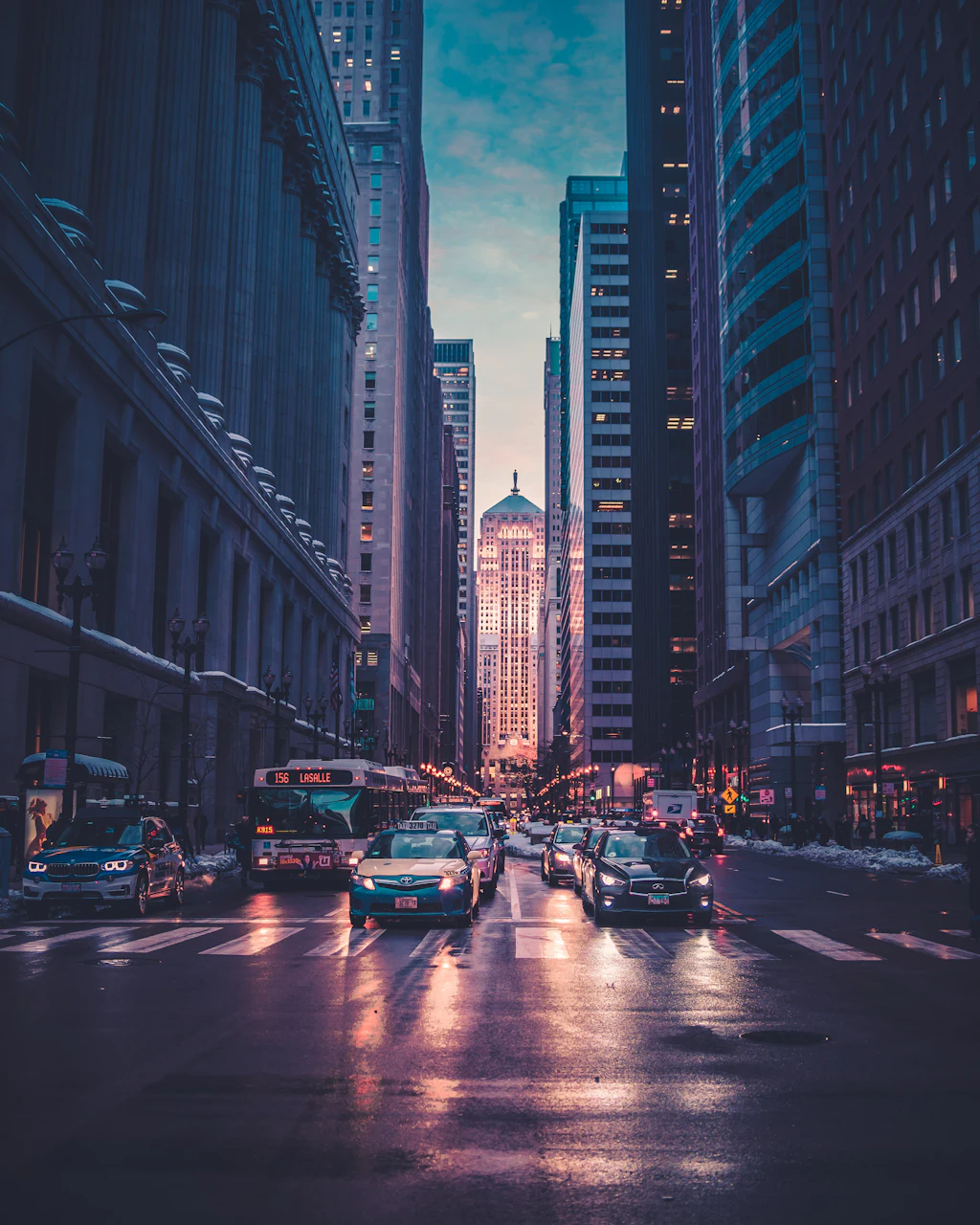 City street at dusk between tall buildings, with traffic lights and headlights below