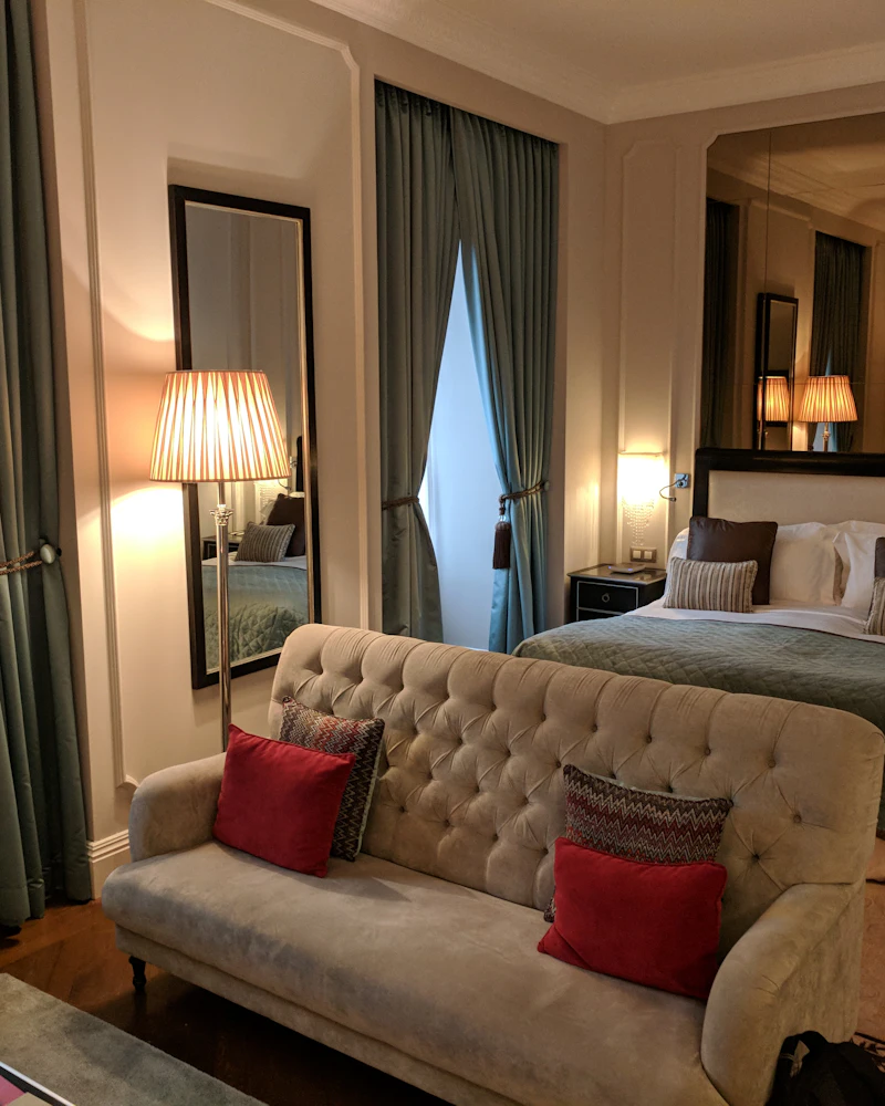 Hotel suite with a tufted sofa at the foot of a large bed, tall curtains and table lamps